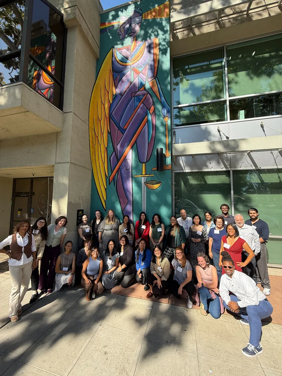 About thirty-five people stand and crouch together in sunlight outside a building, in front of a tall painted mural of a winged figure of Justice holding a set of scales.