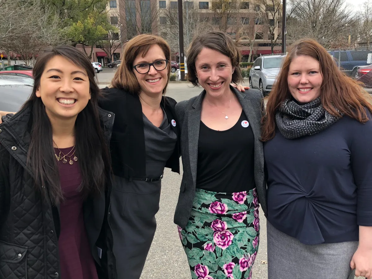 Four women stand close together and smile at the camera on a street, with parked cars and bare trees behind them.