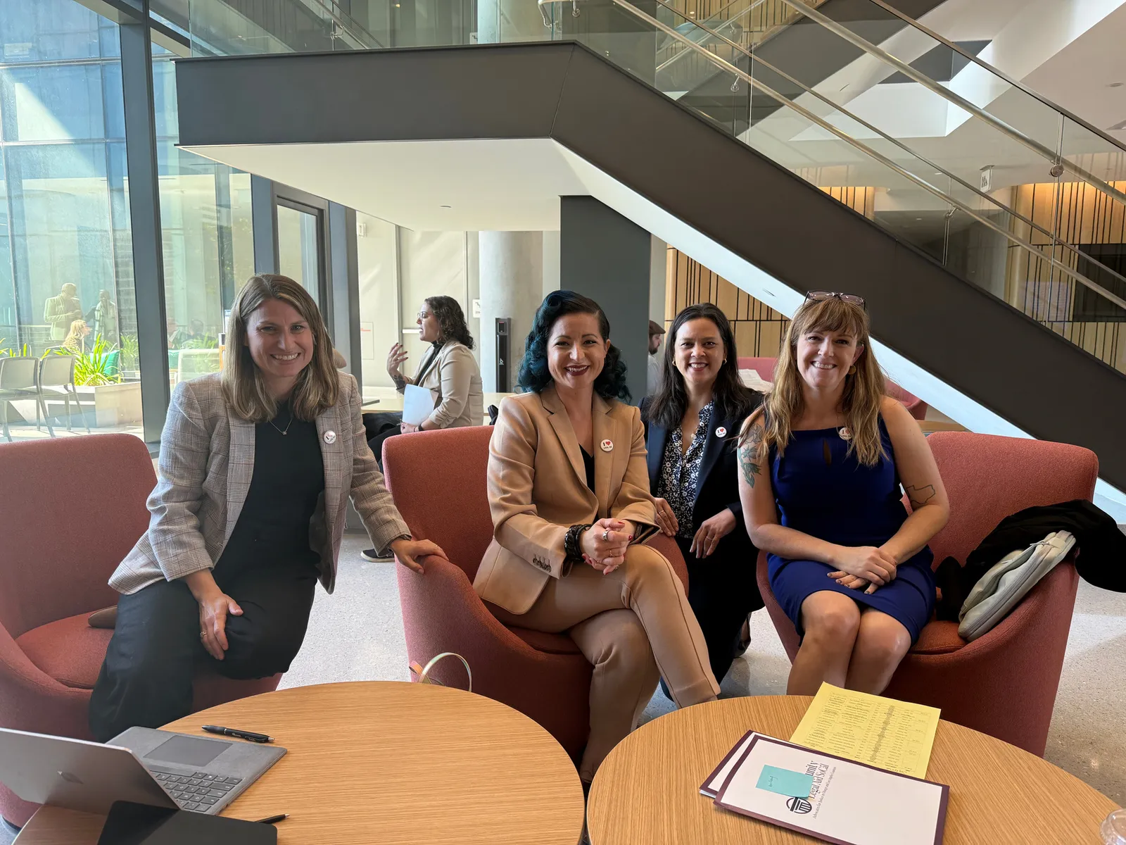 Four people sit together in armchairs in an office lobby, wearing name pins, with a folder and printed schedule on the low table in front of them.