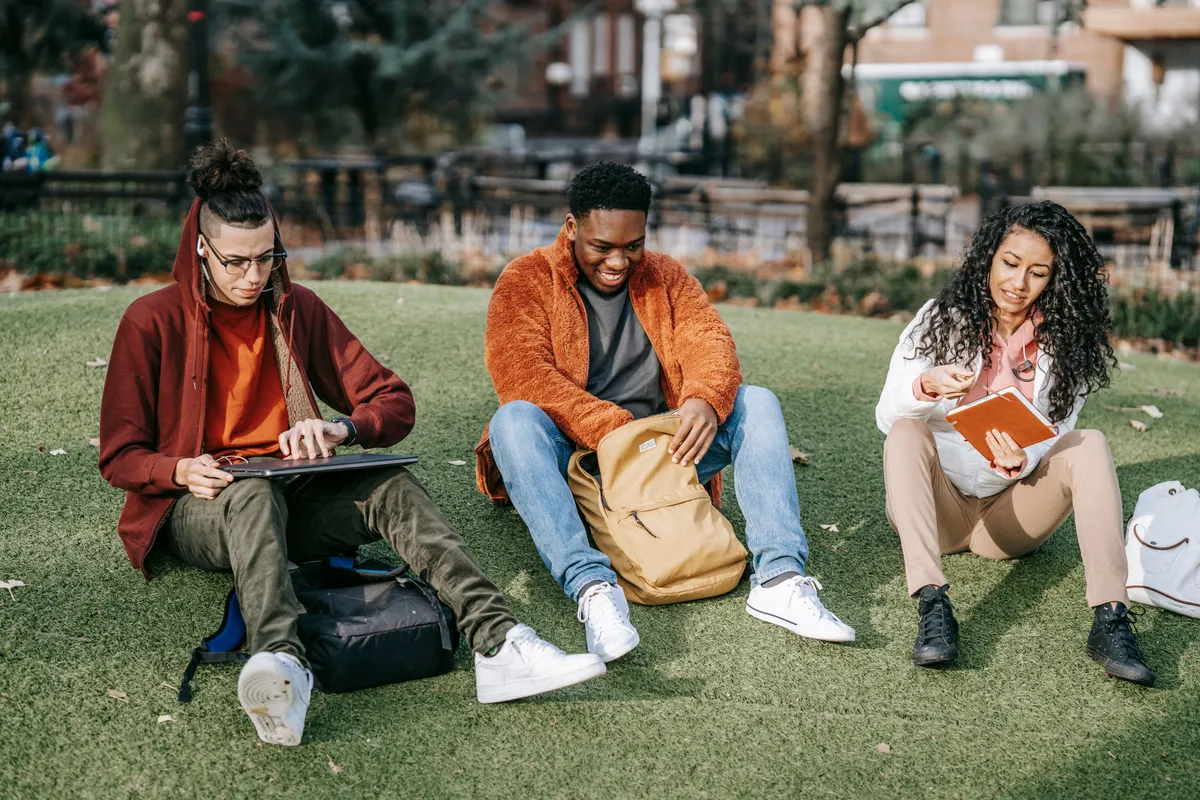 Three students sitting on a campus lawn with a laptop, a notebook and a book.