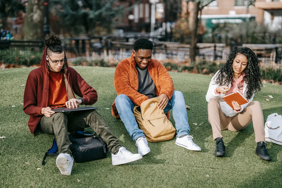 Three students sitting on a campus lawn with a laptop, a notebook and a book.