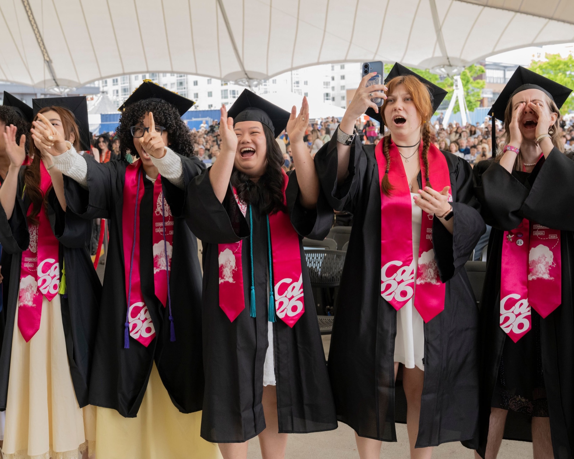 MassArt graduates cheer at an outdoor commencement ceremony.