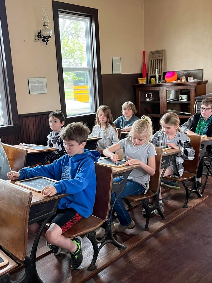 Students in period desks inside the Little Red Schoolhouse, a girl in a blue sweater at the front