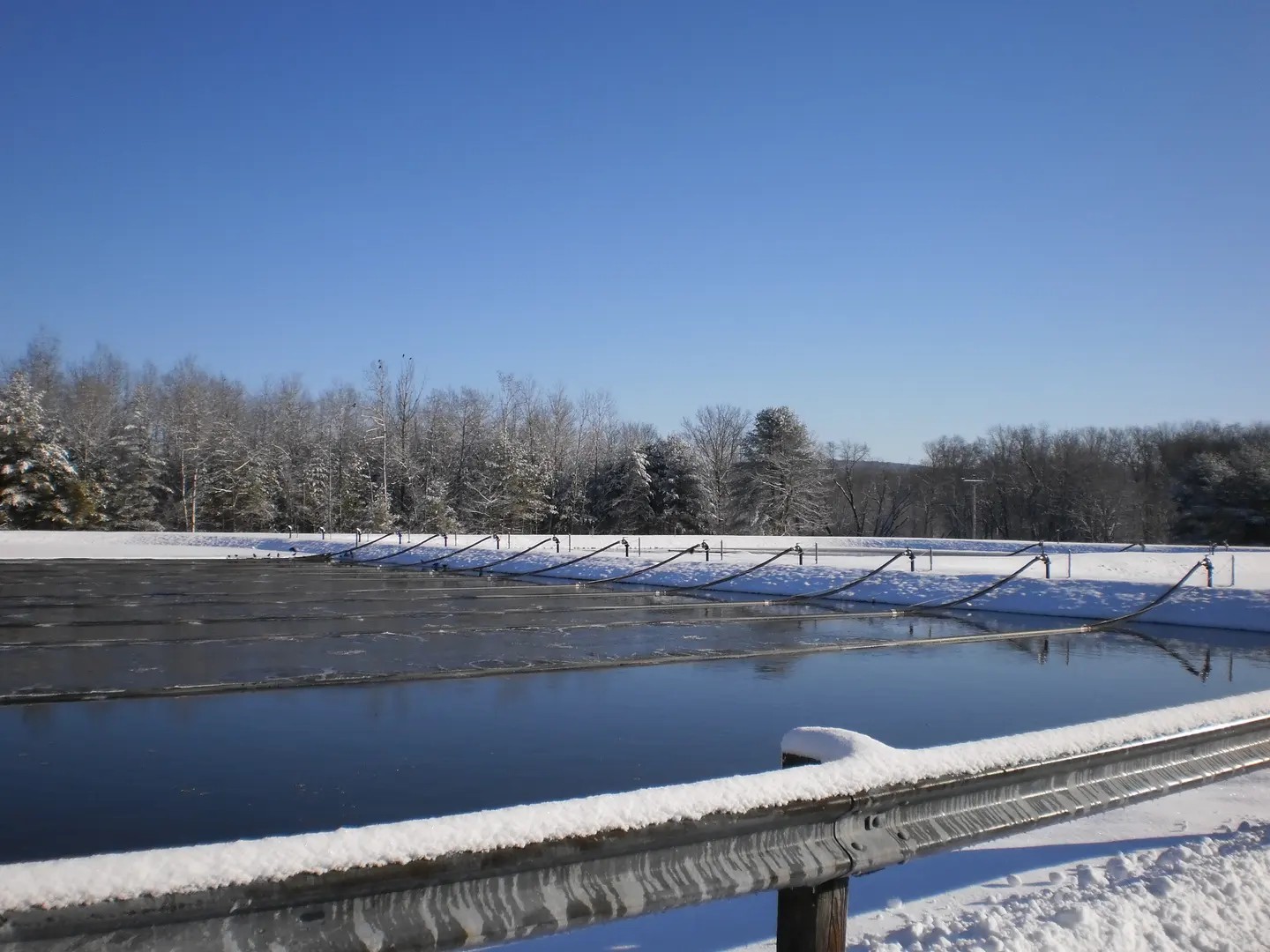 A wastewater lagoon with snow on its banks and bare trees beyond