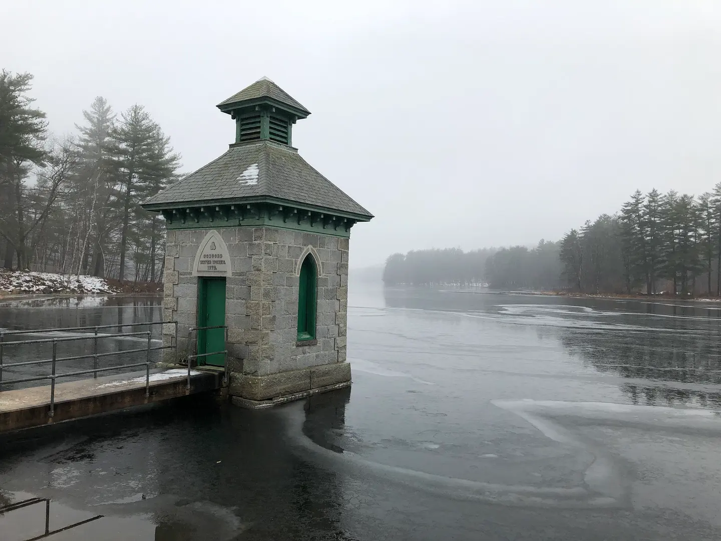 A small brick gatehouse with a cupola standing at the edge of open water in fog
