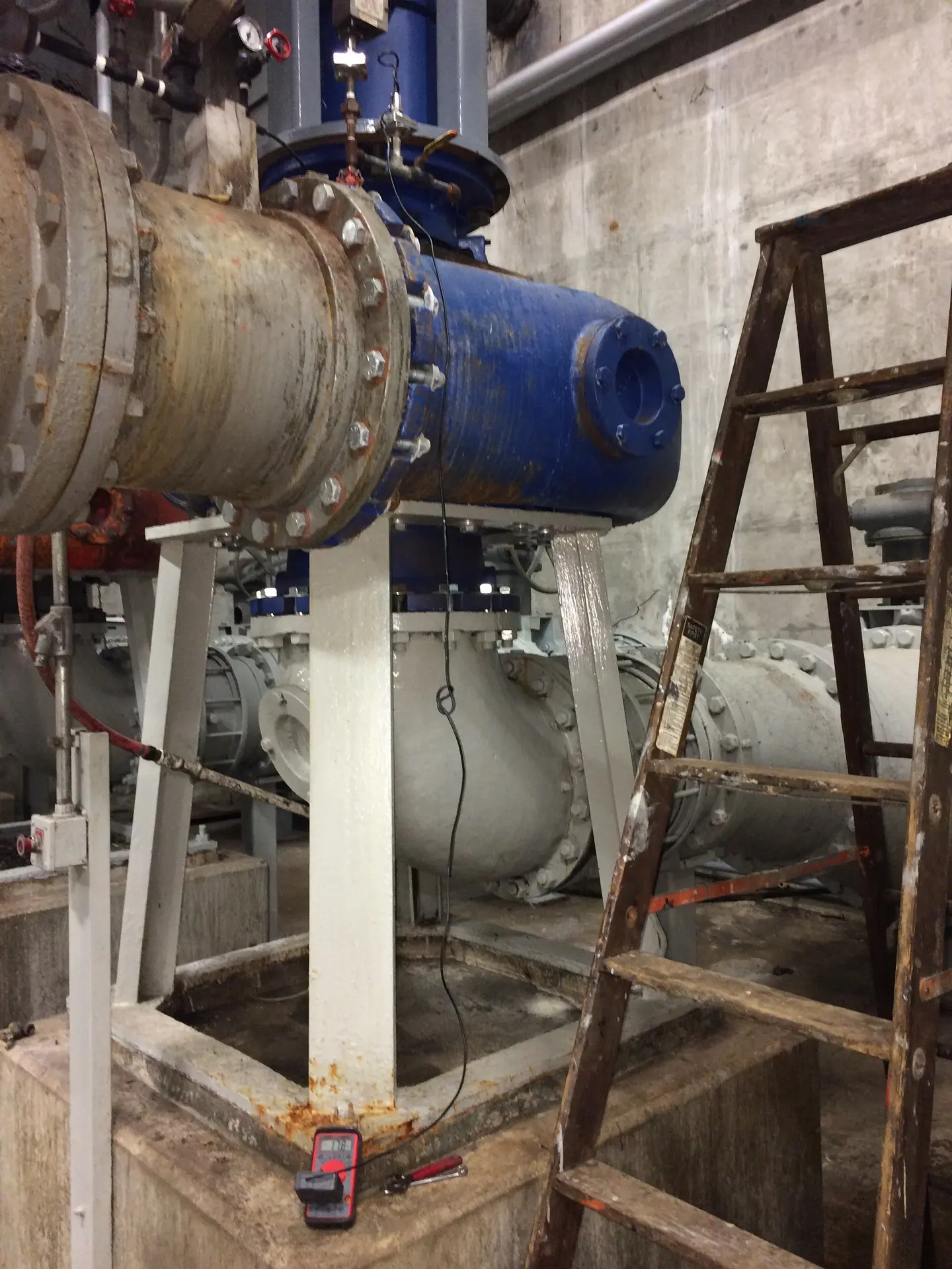 Large blue and gray pipes and a stepladder in a treatment facility basement