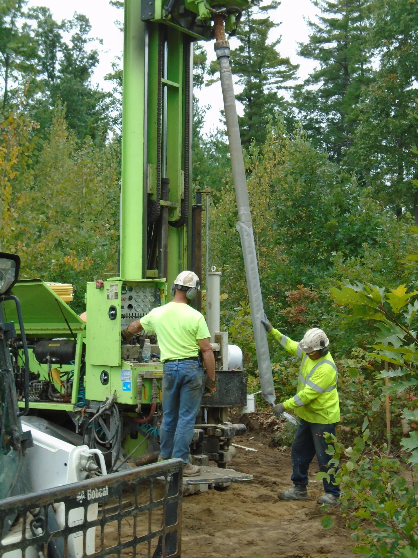 Two workers in safety vests run a green drill rig in the woods