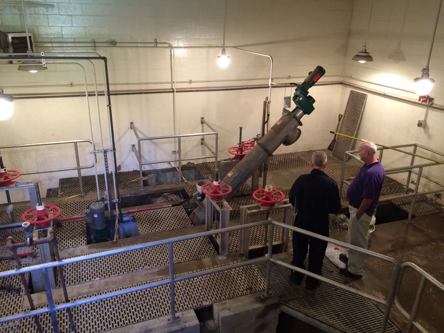 Two men stand looking over screening equipment and red valves inside a treatment facility