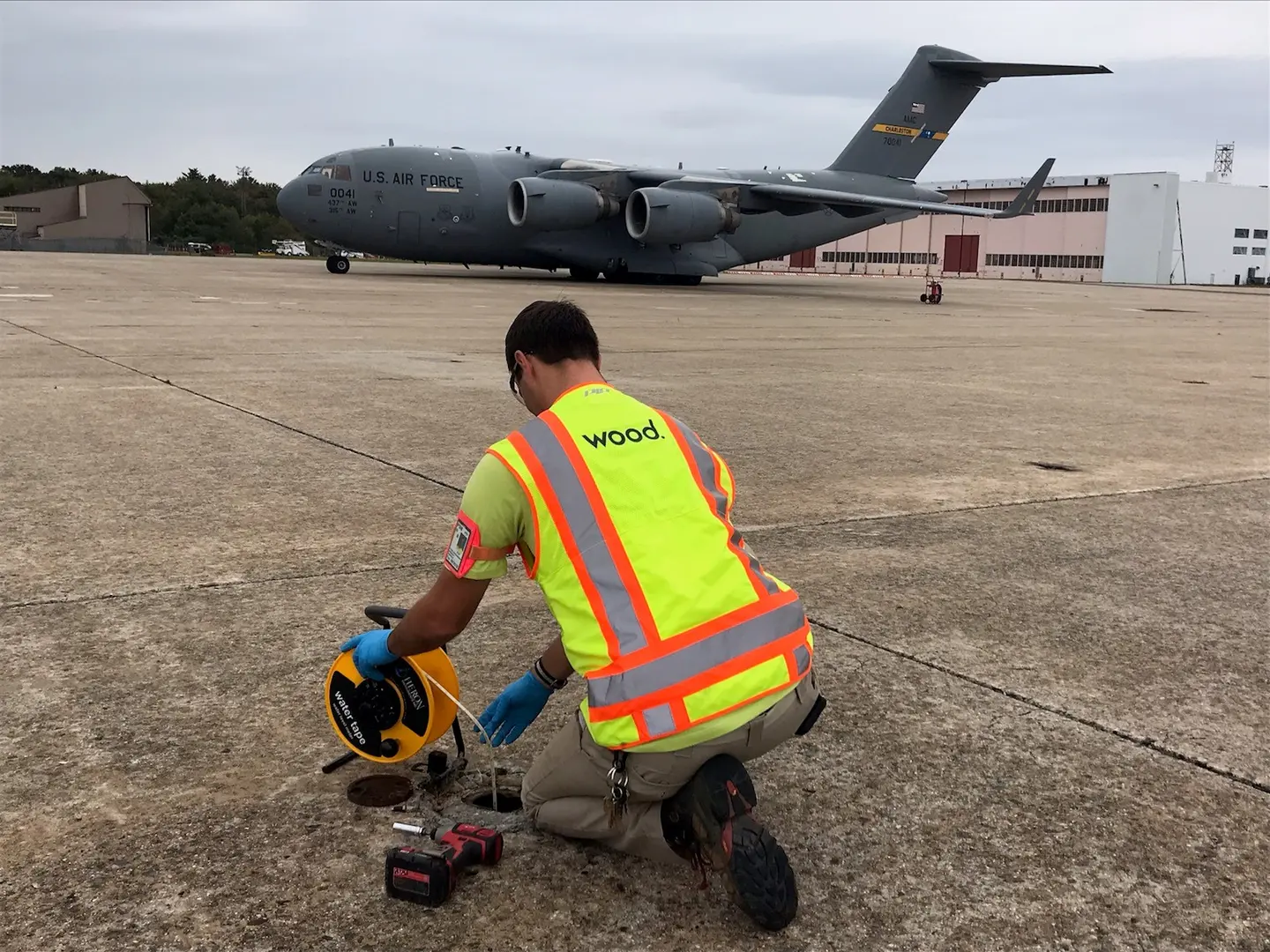 A worker in a safety vest lowers a gauge into a monitoring well on an airfield, a cargo plane behind