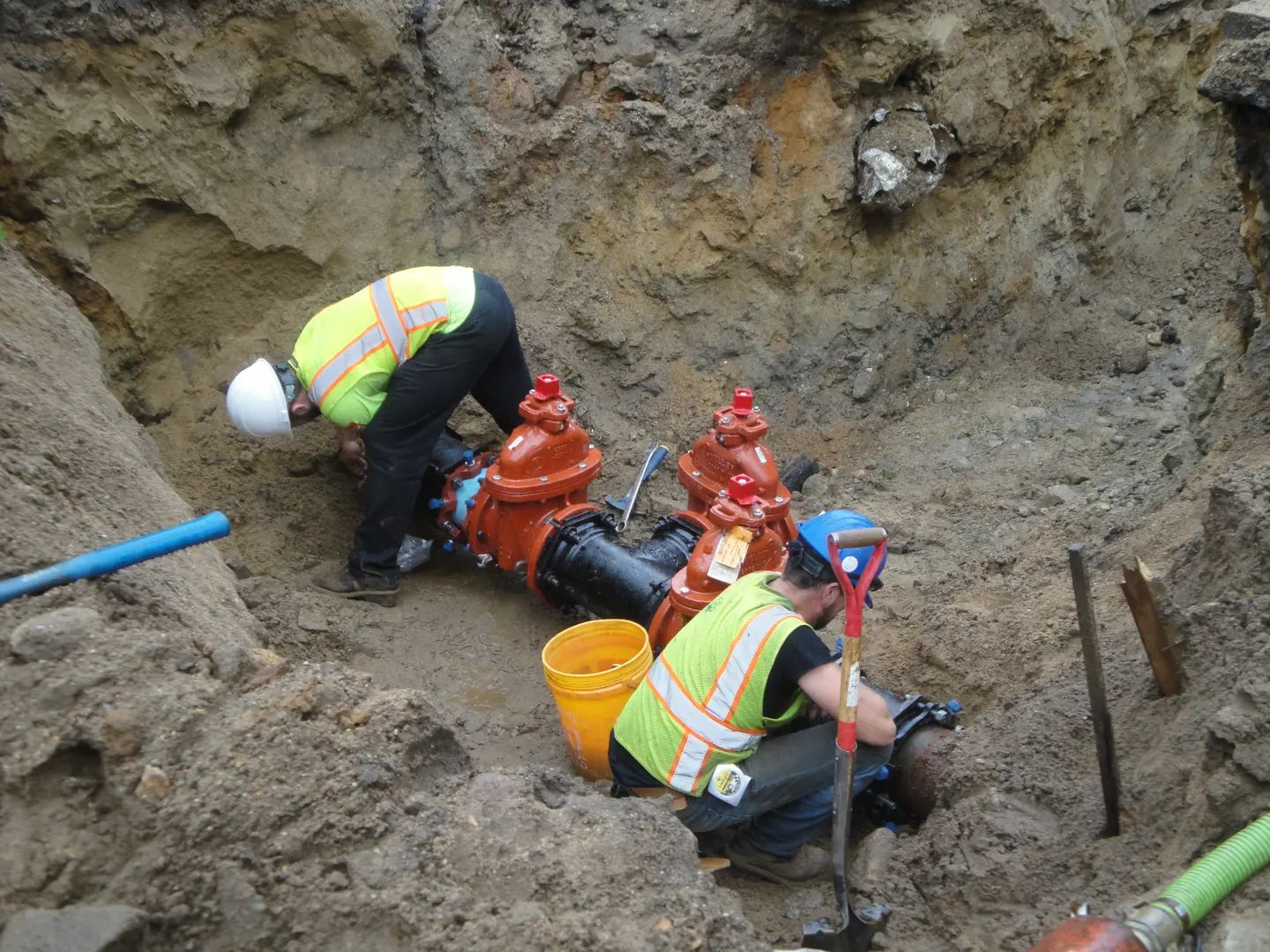 Two workers in hard hats and safety vests set red valves in a sandy trench beside blue pipe