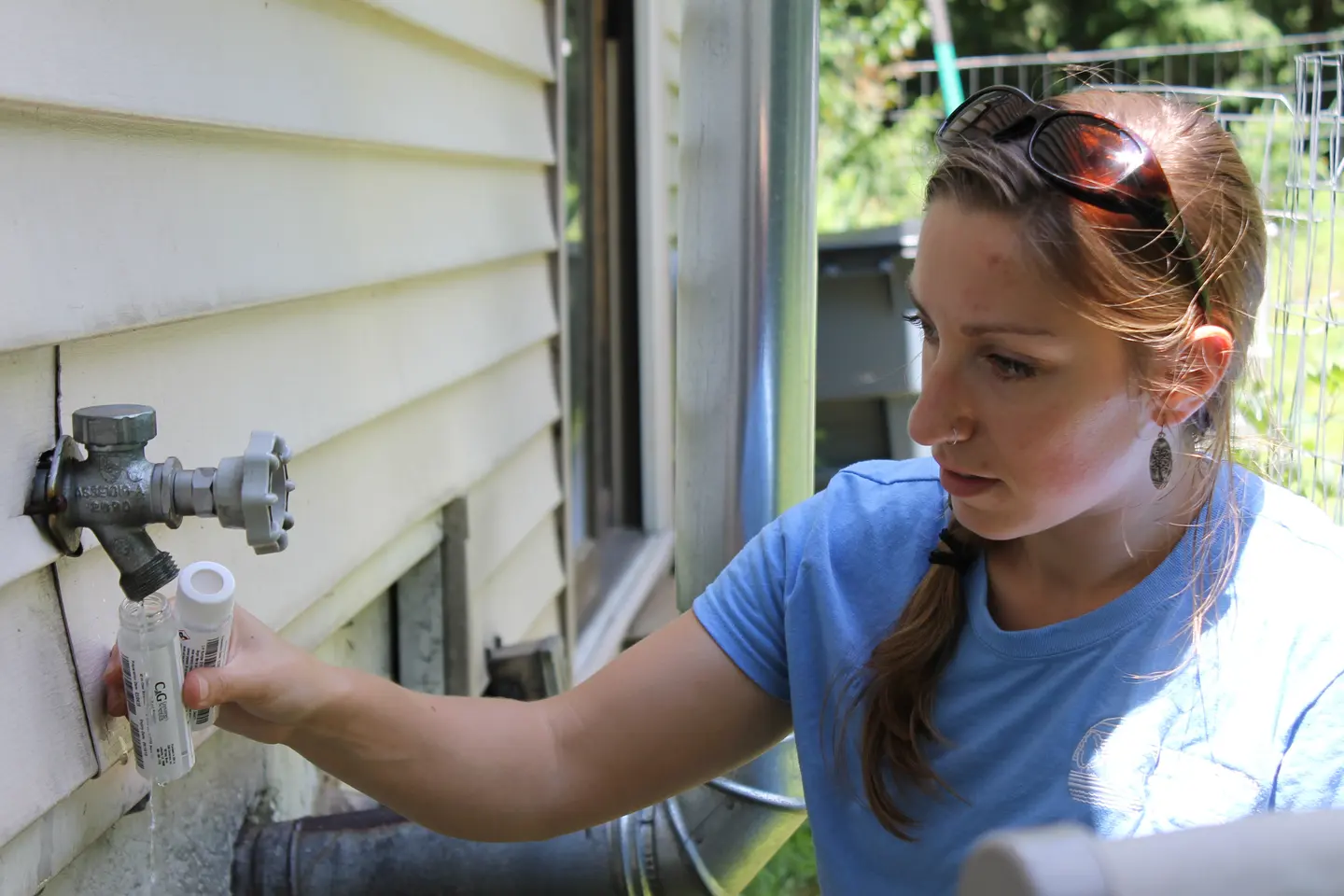 A woman fills a small sample bottle from a spigot on the side of a house