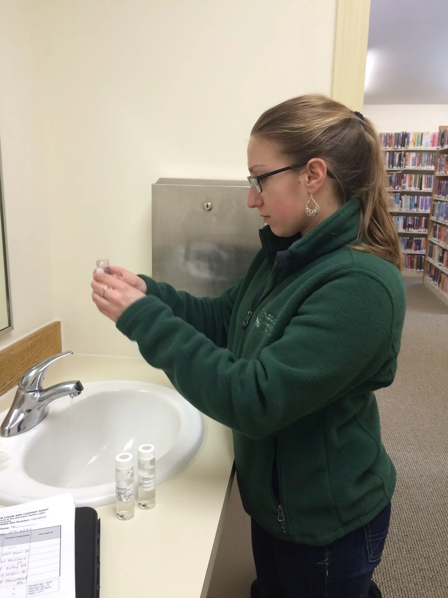 A woman in a green fleece holds up a sample bottle beside a sink