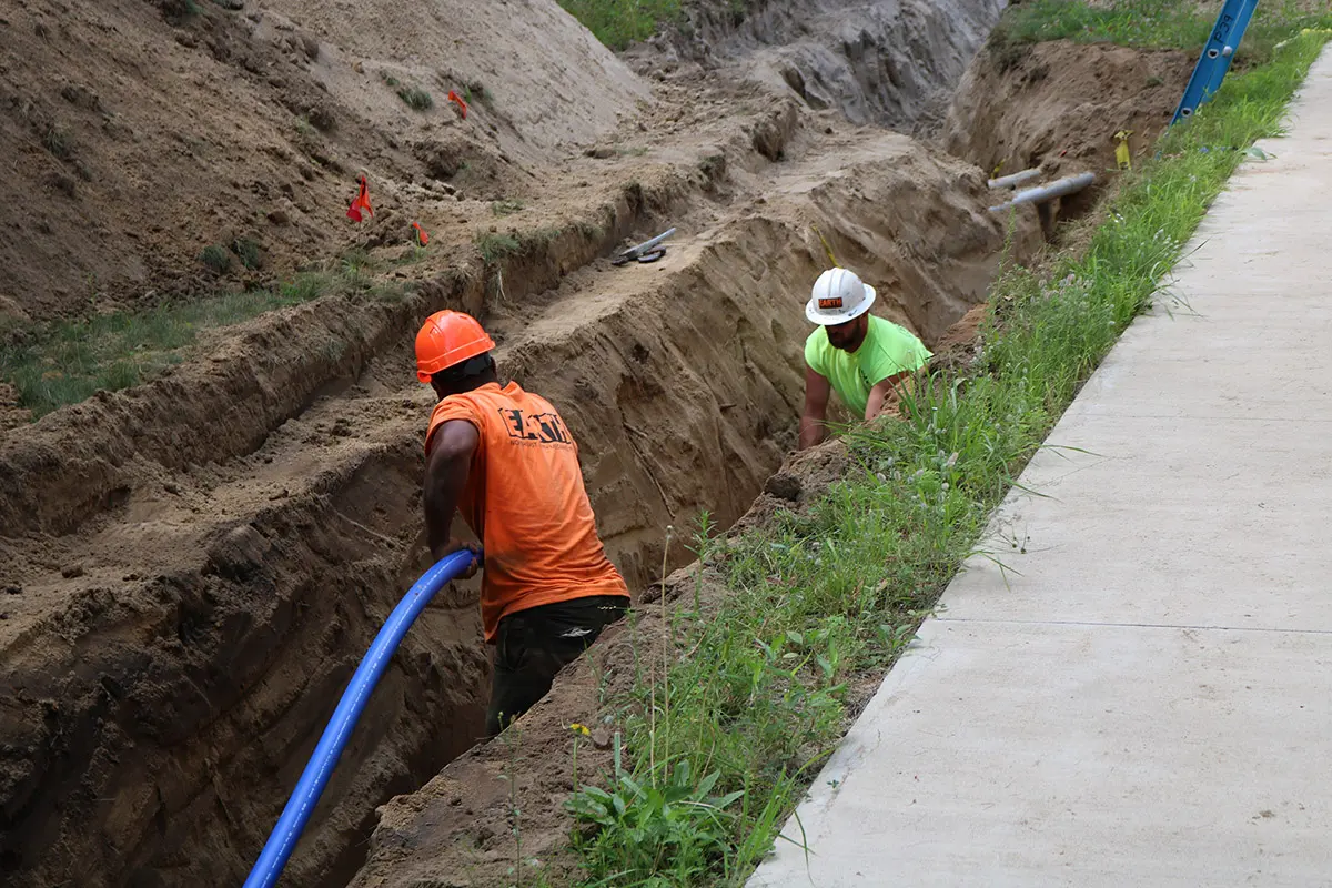 Two workers lay blue water pipe in a long trench beside a sidewalk