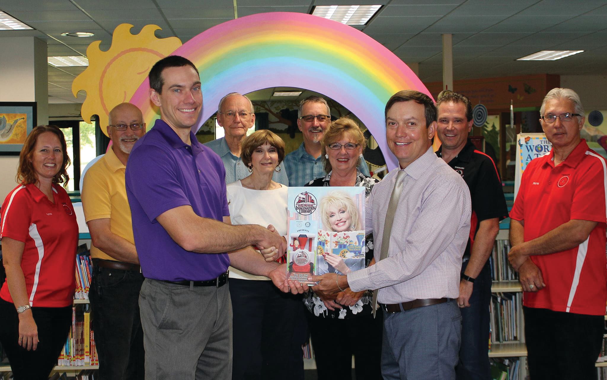 People in Onward! red polos with partners holding a Dolly Parton's Imagination Library poster in a library children's area
