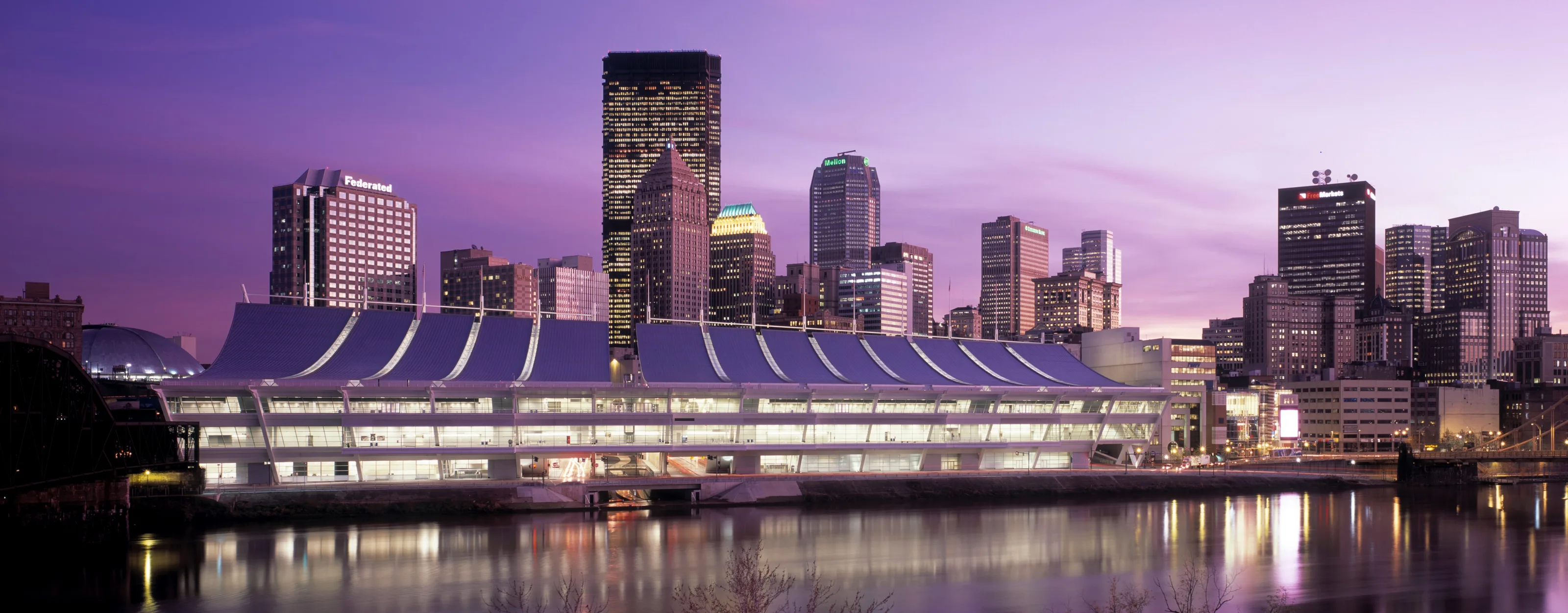 The David L. Lawrence Convention Center at dusk, seen across the Allegheny River with the downtown skyline behind it.