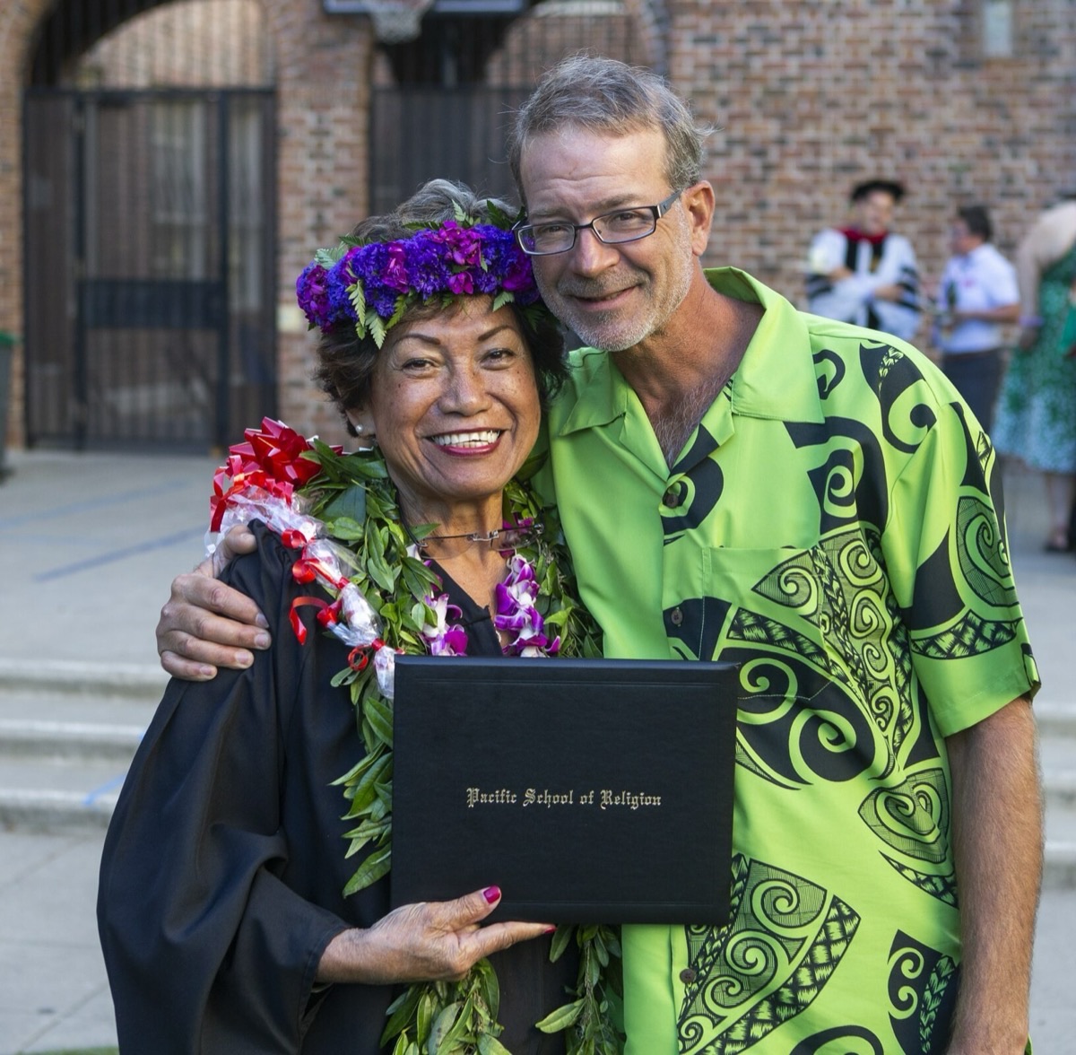 A graduate embraces family outside a brick campus building, diploma folder in hand