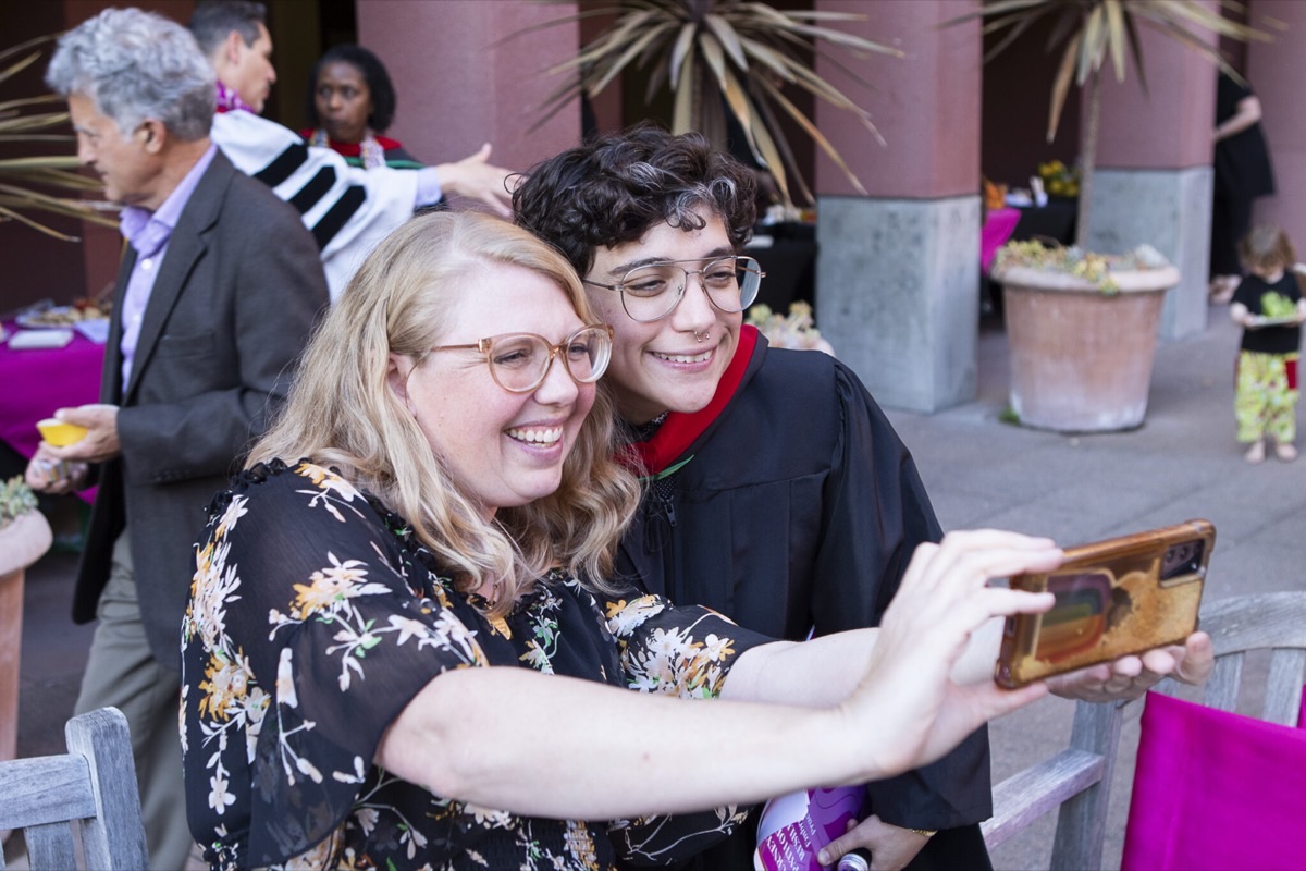 A graduate in regalia takes a selfie with a guest on a sunny campus courtyard