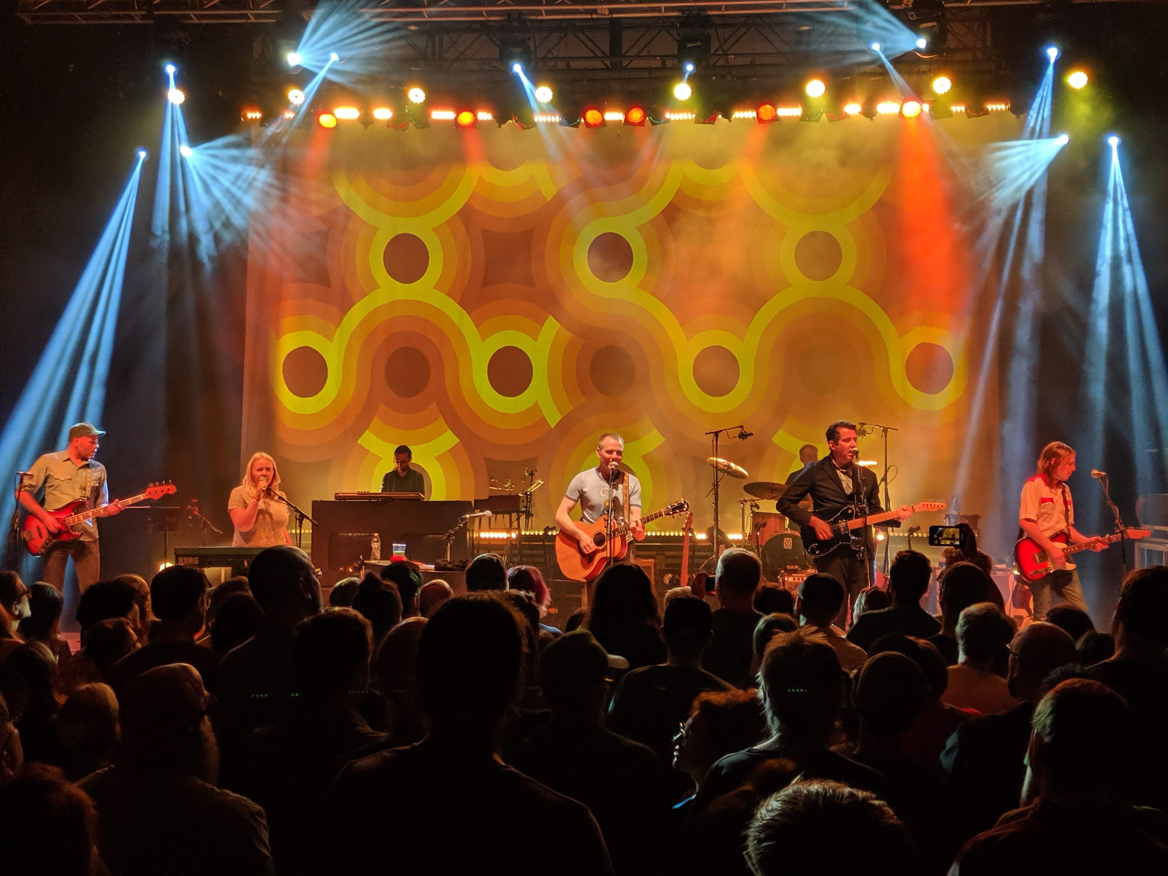 A band performing under warm stage lights to a packed standing crowd inside the Royal Oak Music Theatre