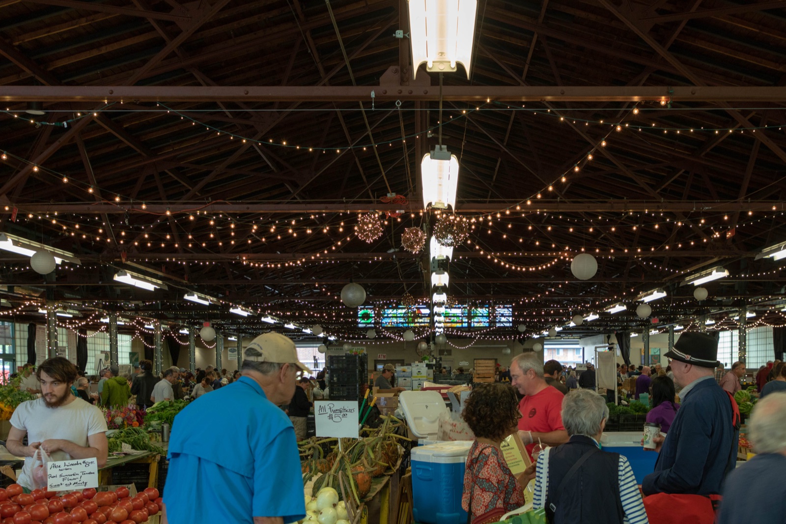 Shoppers and vendors under strings of warm lights inside the Royal Oak Farmers Market hall