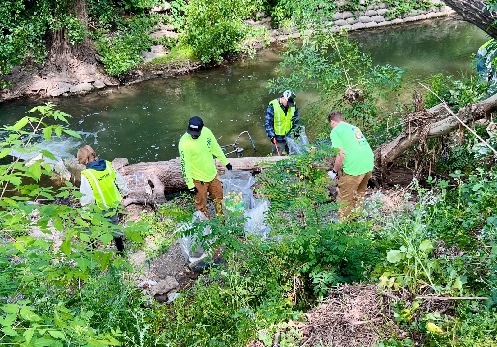 Four Valley Water cleanup workers in high-visibility clothing clearing debris and a fallen log from a creek channel