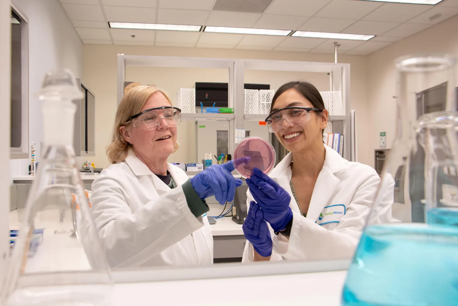Two Valley Water laboratory scientists in lab coats, safety glasses and gloves examining a sample dish together