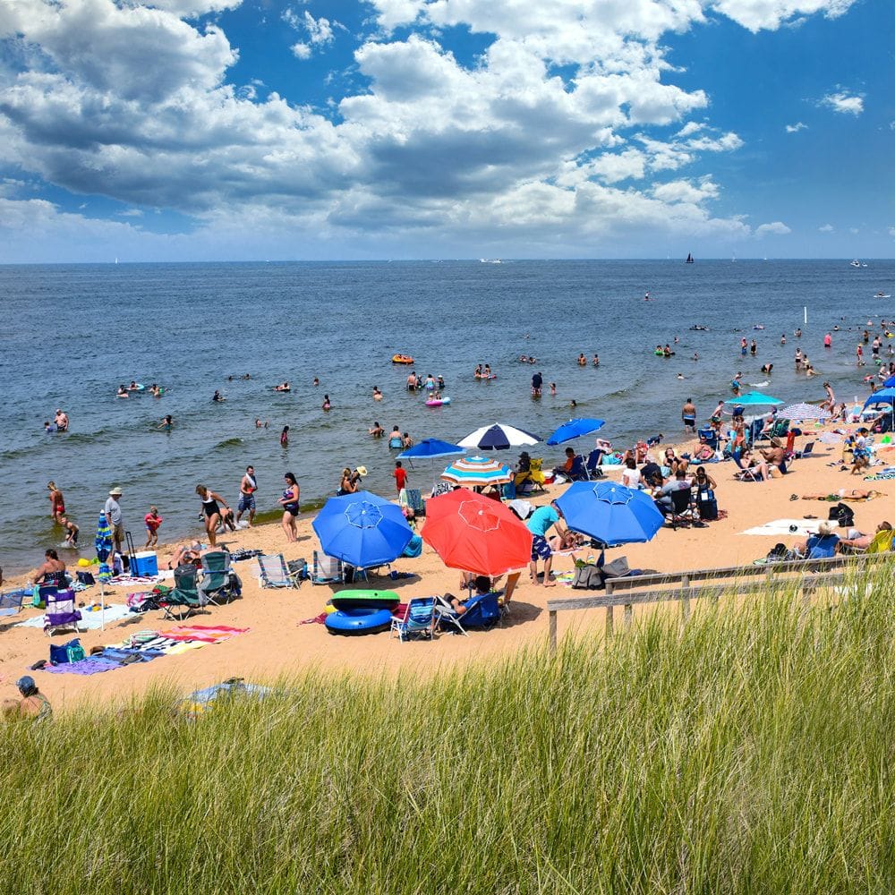 Oval Beach on a summer day, crowded with umbrellas, towels, and swimmers along Lake Michigan, seen past the dune grass.