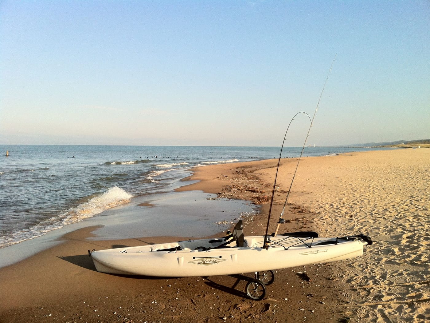 A white fishing kayak with two rods upright rests on the sand at the edge of Lake Michigan, the beach curving away north.