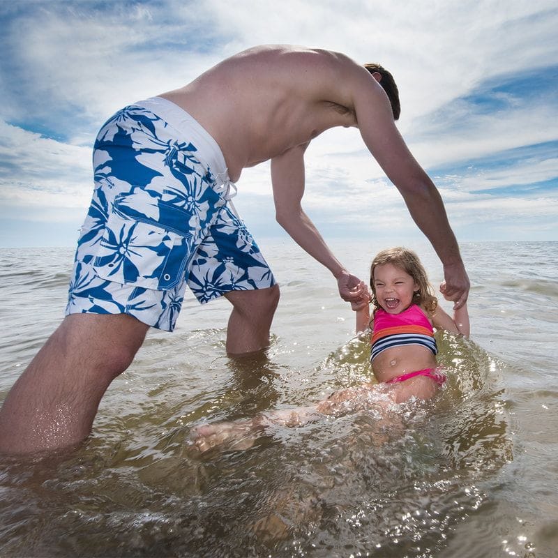 A father bends to hold the hands of his laughing young daughter as she sits in the shallow water of Lake Michigan.