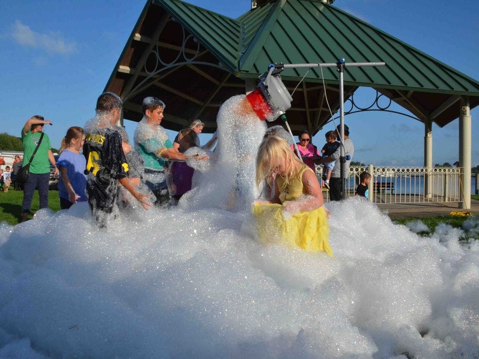 Children and adults stand in deep white foam under the Coghlin Park gazebo roof as a foam cannon pours out more.