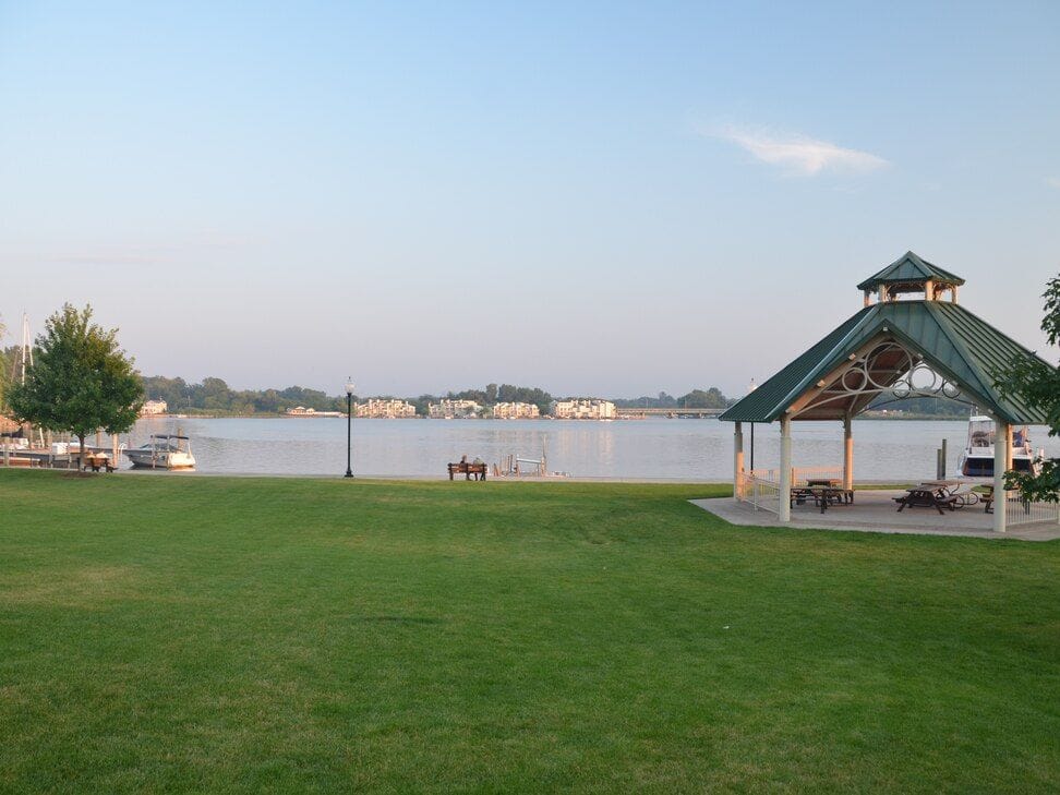 The green-roofed Coghlin Park gazebo at the edge of a wide lawn, with the Kalamazoo River and the far shore behind it.