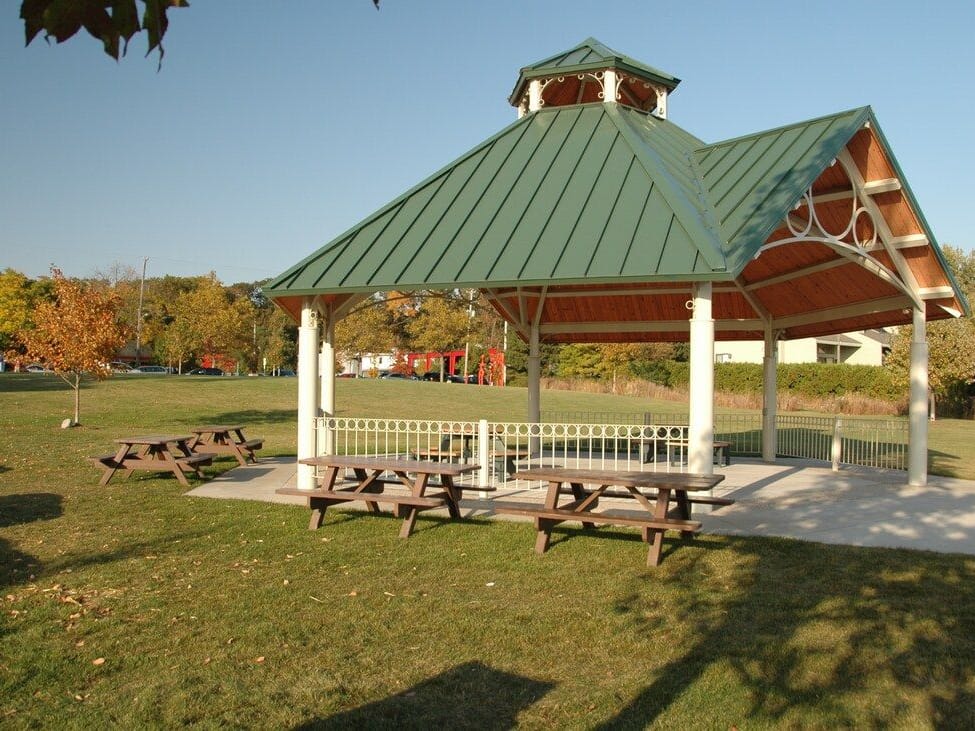 The green-roofed Coghlin Park gazebo with picnic tables under it and on the lawn beside it, a young maple turning orange behind.