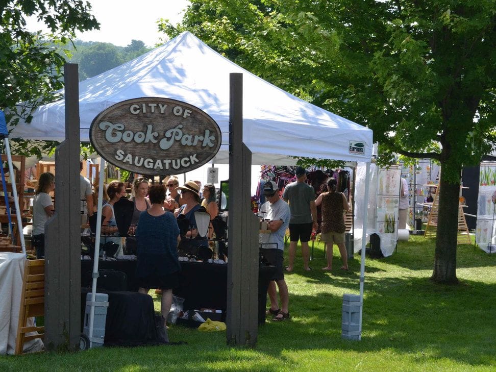 Shoppers crowd an art fair tent on the lawn beside the carved City of Saugatuck Cook Park sign, under shade trees.