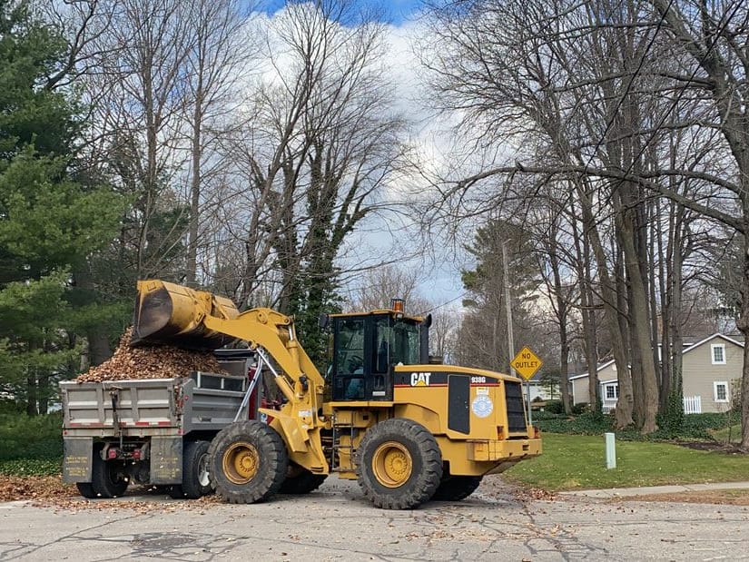 A yellow front loader with the City seal on its side tips a bucket of leaves into a dump truck on a residential street, under bare trees and tall pines, beside a No Outlet sign.