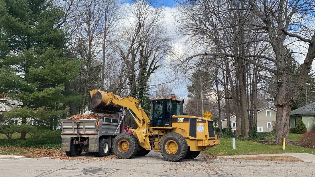 A Public Works front loader lifts leaves into a dump truck on a residential street lined with bare trees.