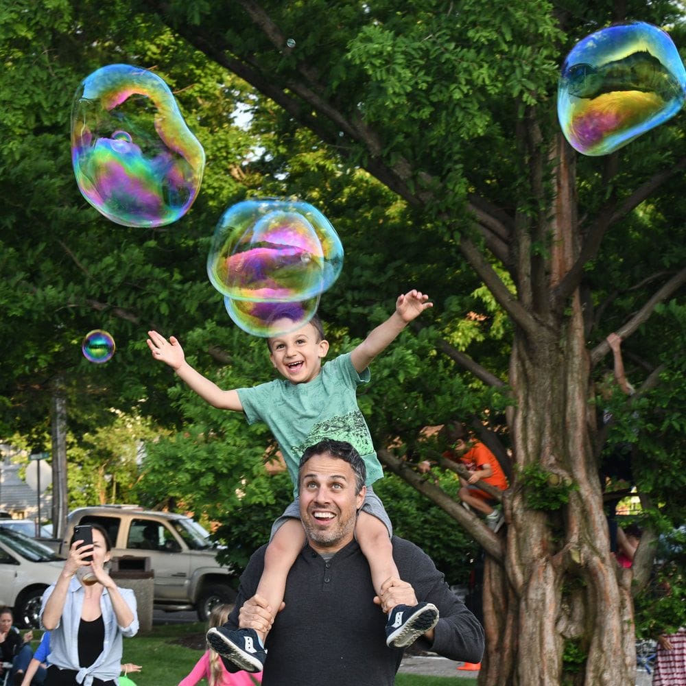 A laughing boy on a man's shoulders reaches up for giant soap bubbles floating under a tree, as people around them take pictures.