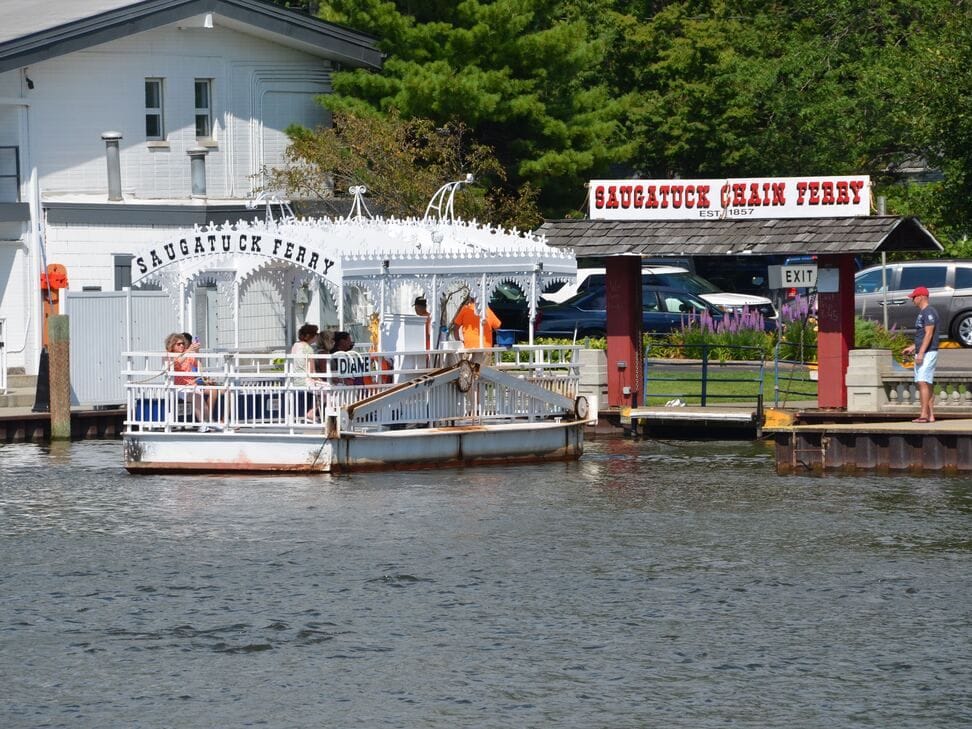 The white Saugatuck Chain Ferry at its landing with passengers aboard, under the red Saugatuck Chain Ferry sign.