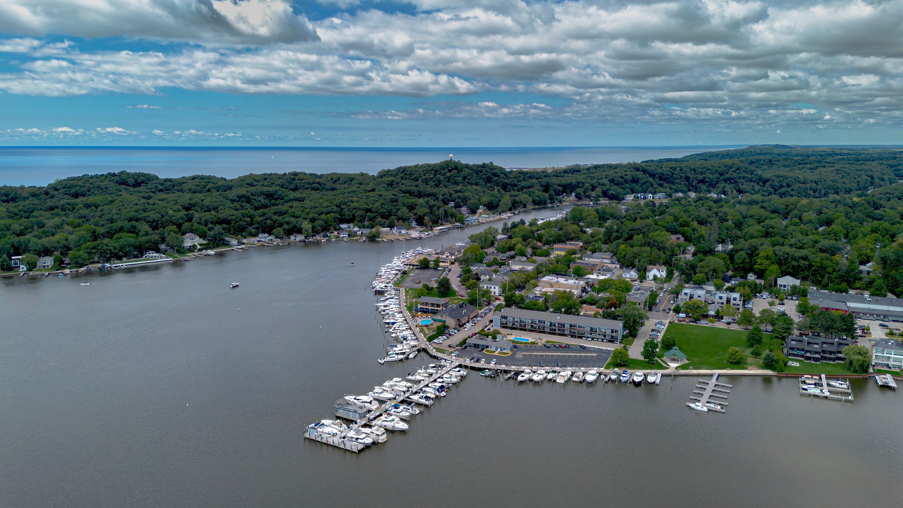 Aerial view over Kalamazoo Lake of the marina docks and downtown Saugatuck, with the wooded dunes and Lake Michigan on the horizon.