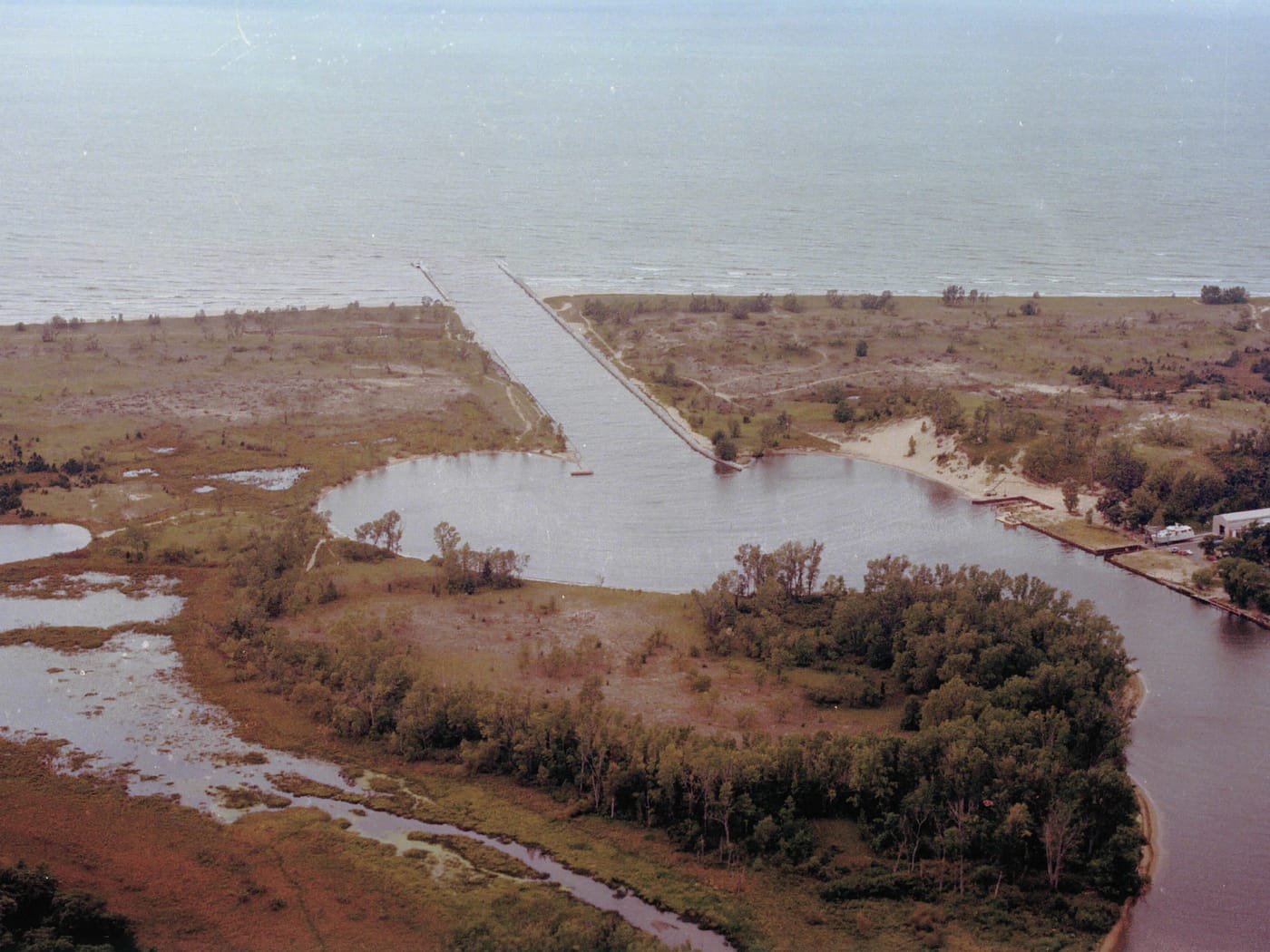 An aerial photograph from 1982 of the river's straight channel running between two piers into Lake Michigan, with marsh, wooded dunes, and the curving river on either side.