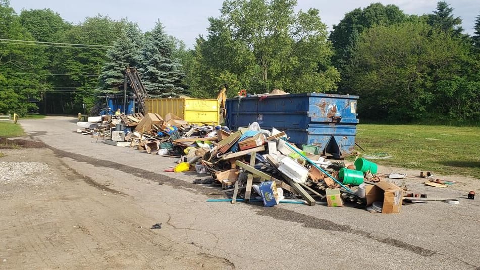 A heap of old furniture, boards, buckets, and boxes on the pavement beside blue and yellow roll-off dumpsters, with evergreens behind.