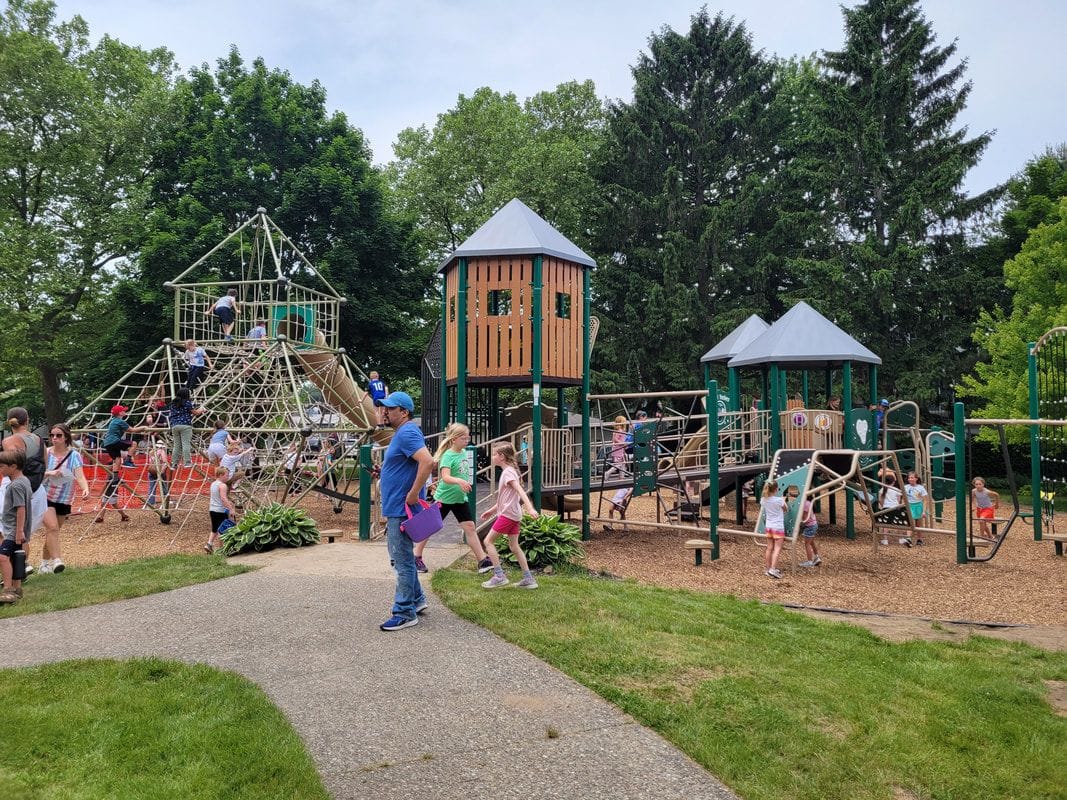 Children climb and run across the new playground at Village Square Park, with tall pines behind.