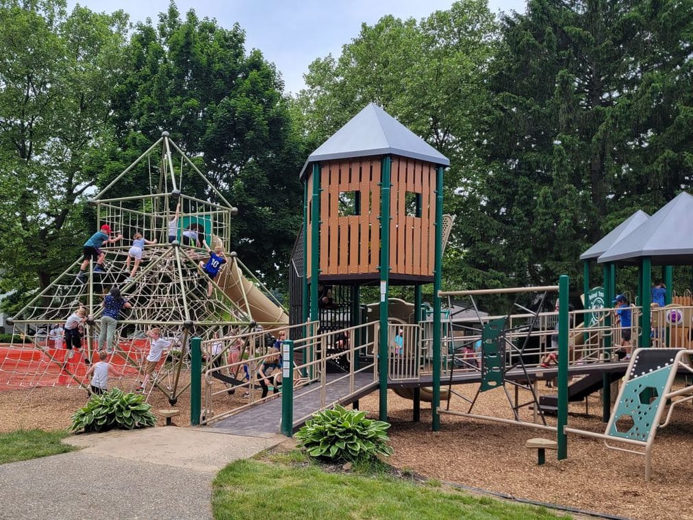 Children climb the rope pyramid and play towers of the new Village Square playground under tall trees.