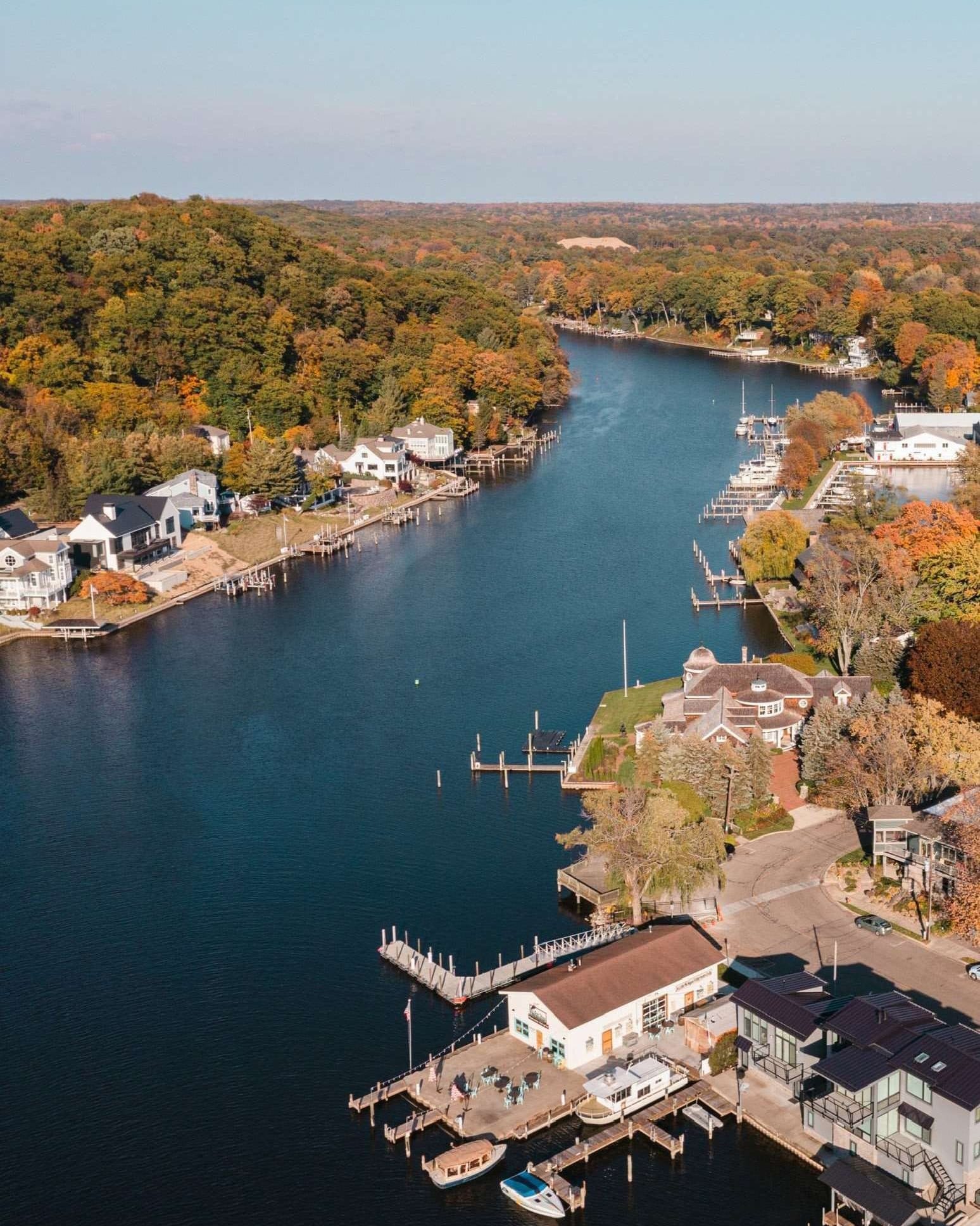 Aerial view of the Kalamazoo River in fall, docks and white boathouses along the near bank and hills of orange and yellow trees beyond.