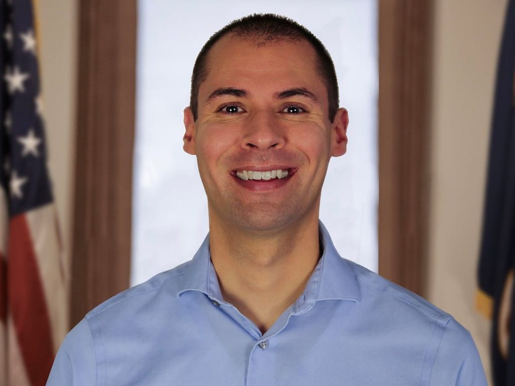 City Manager Ryan Cummins, smiling, in a light blue shirt, with flags behind him.