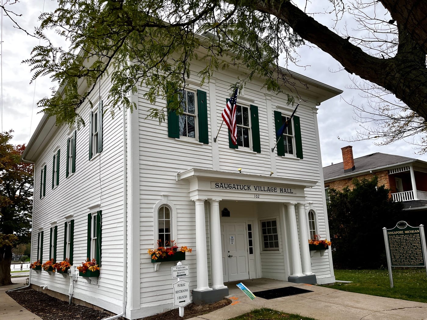 Saugatuck's Village Hall, a two-story white clapboard building with green shutters, a columned porch, and flower boxes, under a large tree.