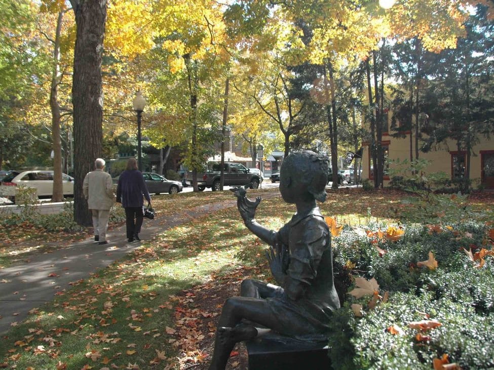 Two people walk a path through Village Square Park under fall color, past a bronze figure seated in the garden bed.