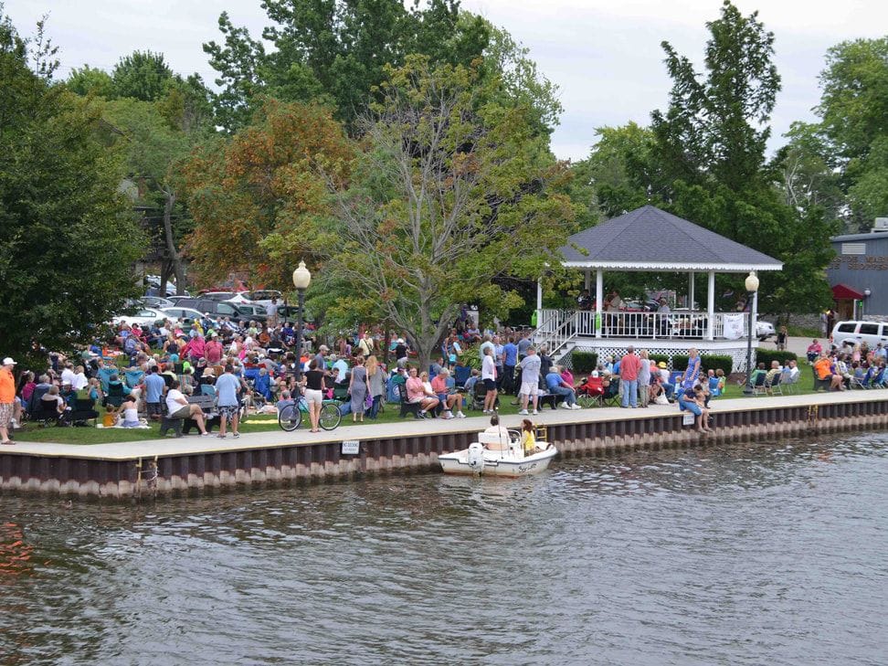 A crowd fills the Wicks Park lawn around the white gazebo along the river seawall, a small boat passing in front.