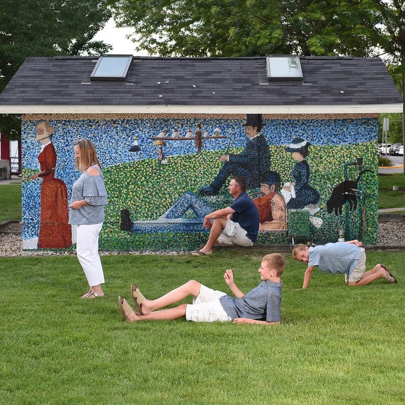 Two boys sprawl on the grass and a woman stands in front of the restroom building's mural of Seurat's A Sunday on La Grande Jatte.