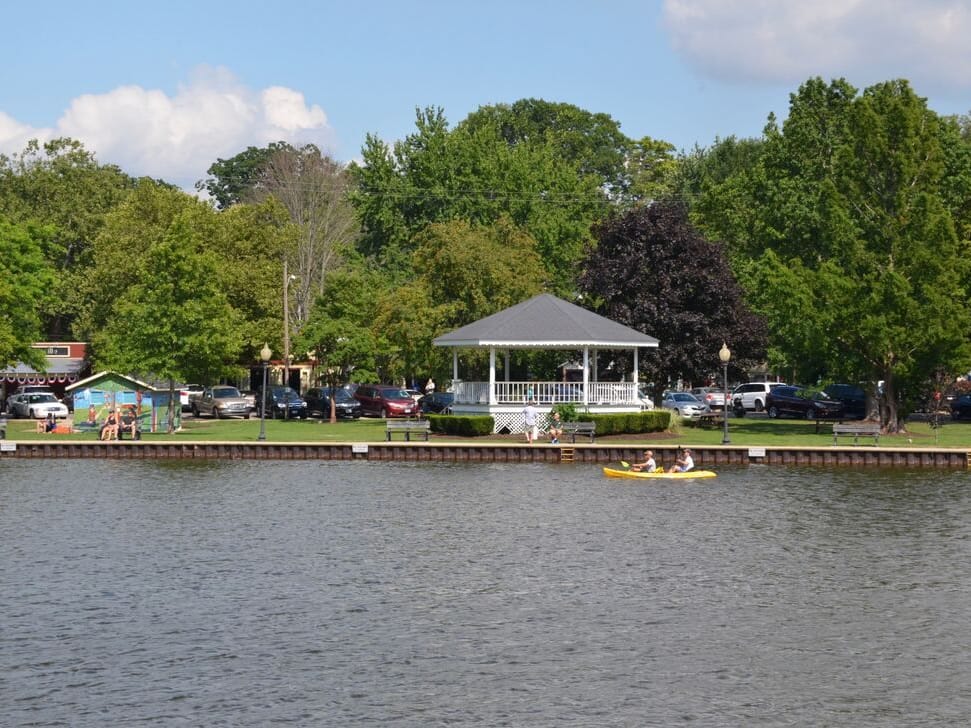 Two people paddle a yellow kayak past the Wicks Park seawall, with the white gazebo, the lawn, and parked cars beyond under tall trees.