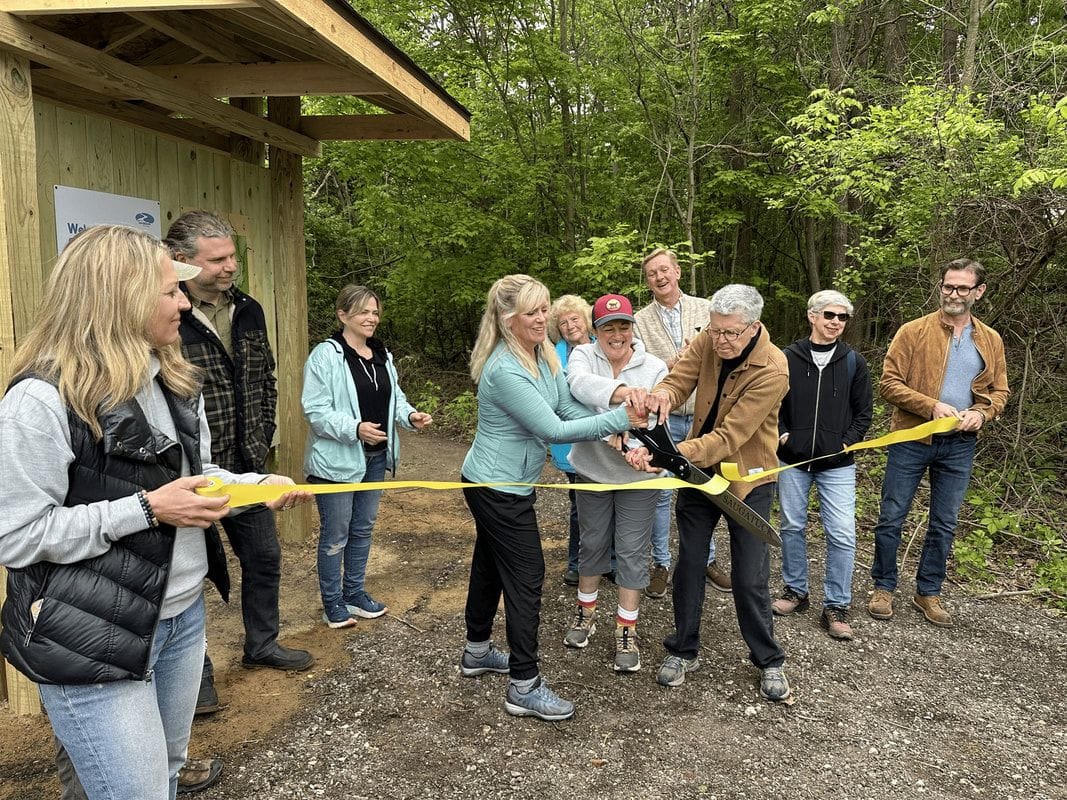 A group of people in fall jackets cut a yellow ribbon together at the trailhead kiosk of the Saugatuck Wilds Nature Preserve.