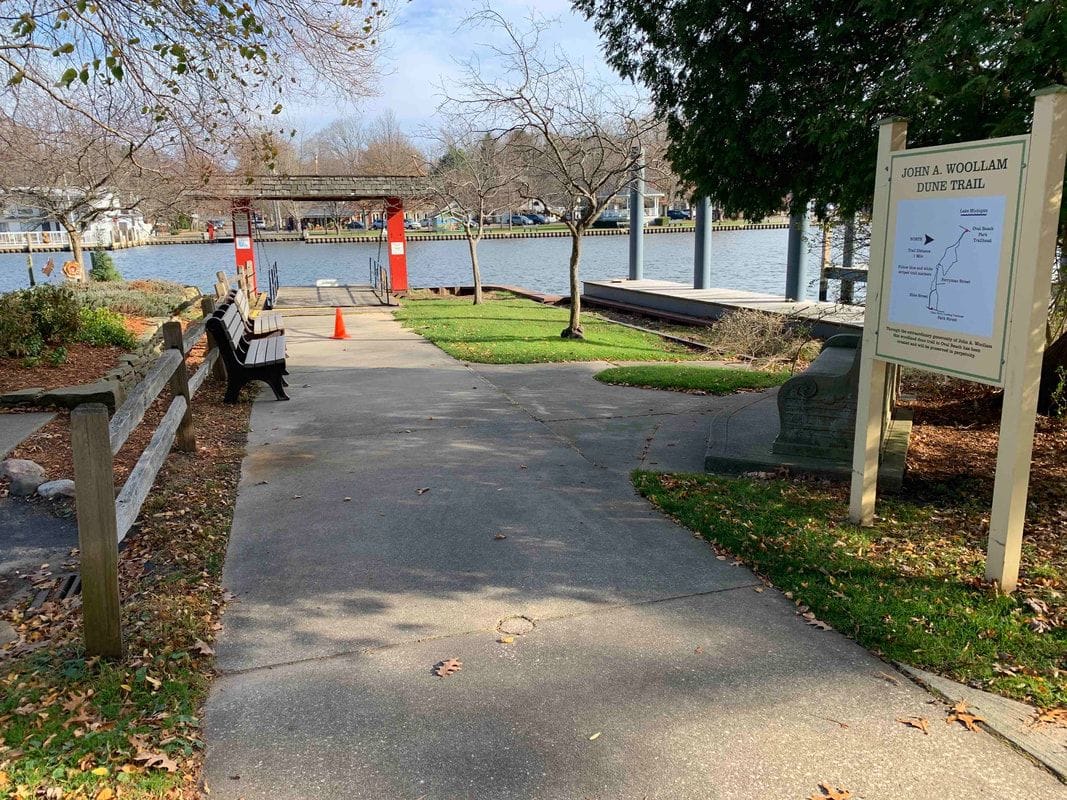 A paved path by the river passes a bench and the trail's signboard and map, with a dock and the red ferry landing sign beyond.
